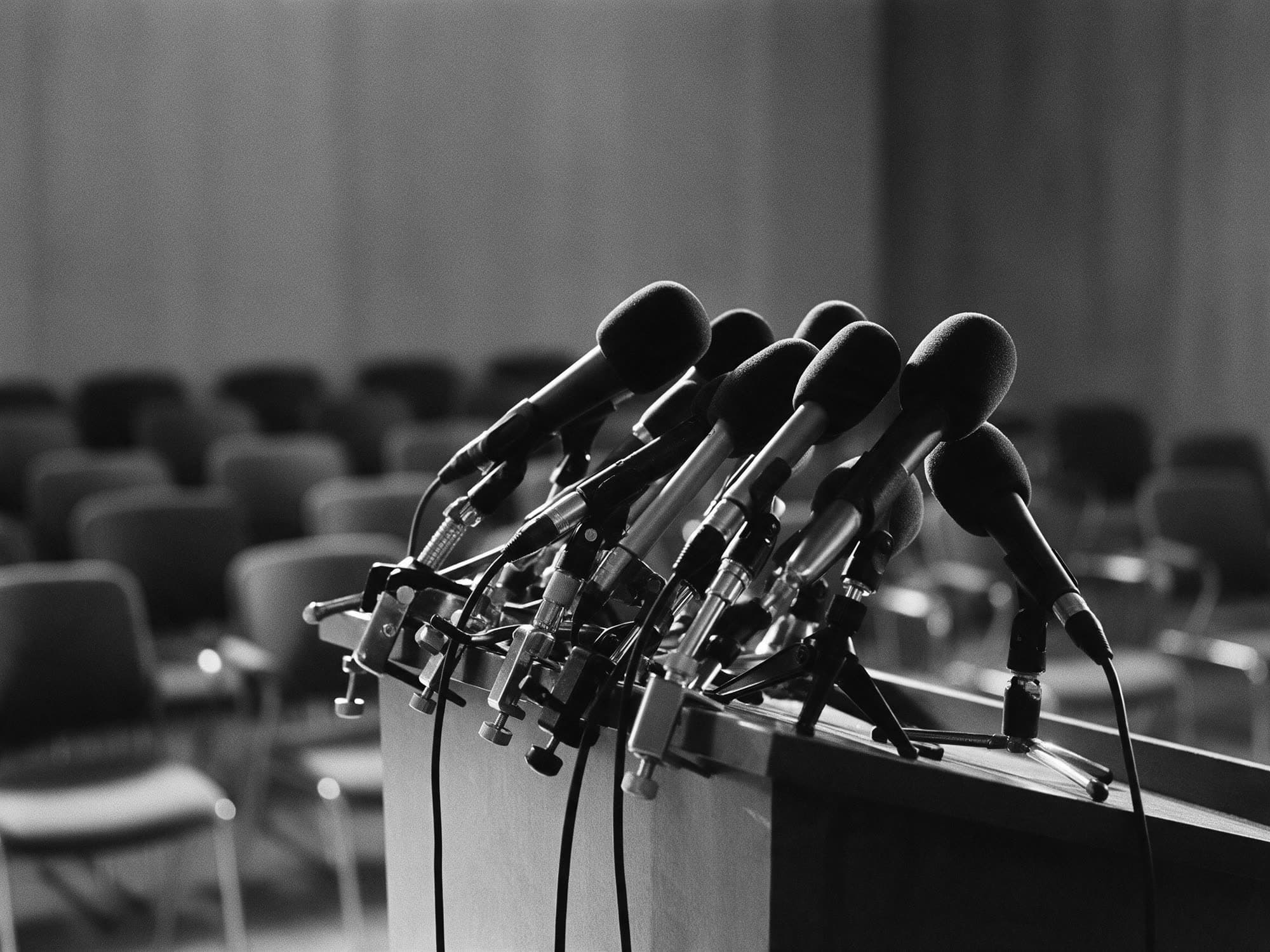 A cluster of press microphones on a podium in an empty room with rows of chairs