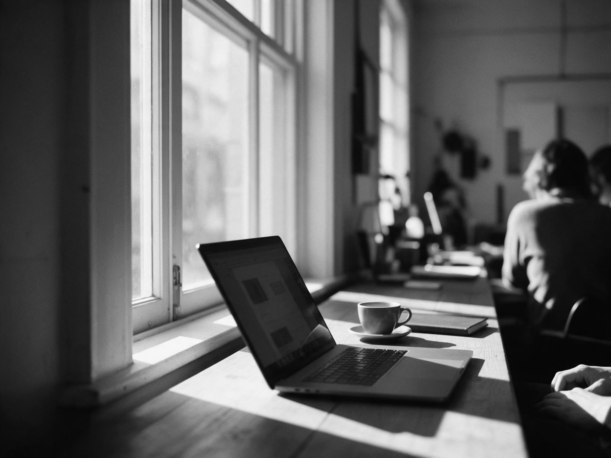 An open laptop and a cup of coffee on a table by a tall window in the morning light
